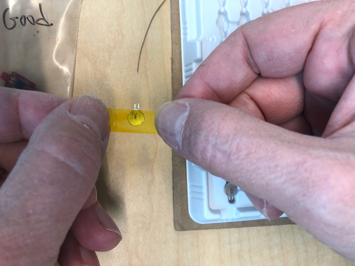 A coin cell held in a strip of Kapton tape before the excess is trimmed